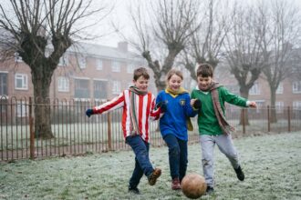Enfants jouant au football dans un parc urbain pendant les vacances de février, portant des maillots colorés et des écharpes
